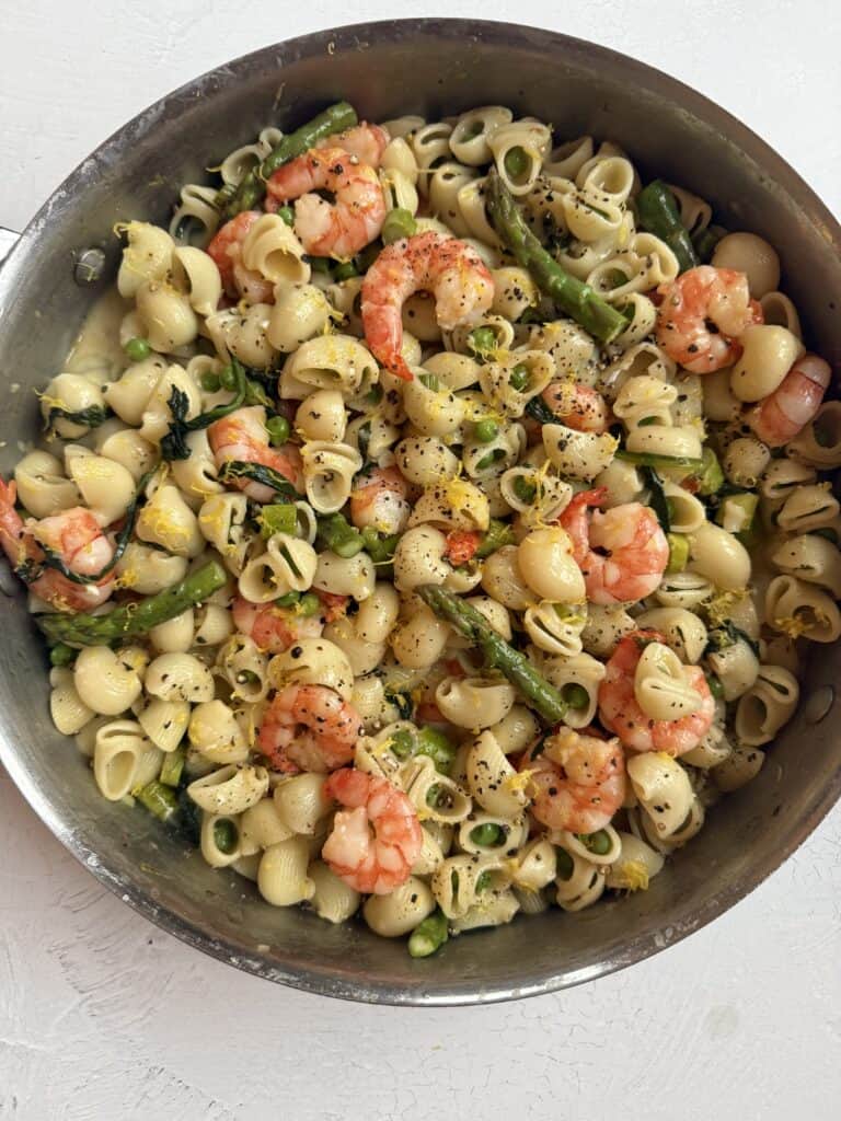 A skillet filled with shell pasta, shrimp, asparagus, and yellow squash, garnished with black pepper and herbs, on a white surface.
