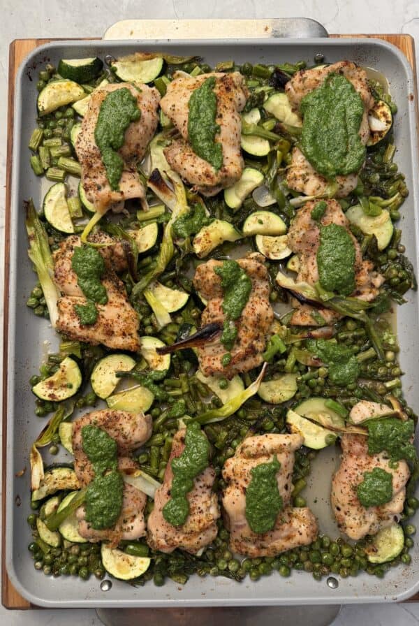A sheet pan with seasoned One Pan Spring Chicken thighs, sliced zucchini, asparagus, green peas, and scallions, all topped with dollops of green herb sauce. The dish is arranged neatly on a gray baking tray.
