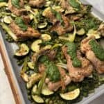 A One Pan Spring Chicken dinner featuring roasted chicken thighs, sliced zucchini, green peas, asparagus, and scallions, all topped with dollops of green herb sauce on a light countertop.