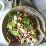 A wooden bowl filled with salad greens, cherry tomatoes, red onion, croutons, and shaved cheese, with two wooden serving spoons. Nearby are bowls of shaved cheese and a pink plate with gold forks.