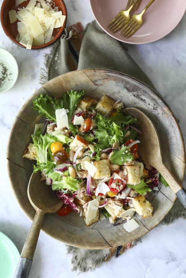 A wooden bowl filled with salad greens, cherry tomatoes, red onion, croutons, and shaved cheese, with two wooden serving spoons. Nearby are bowls of shaved cheese and a pink plate with gold forks.