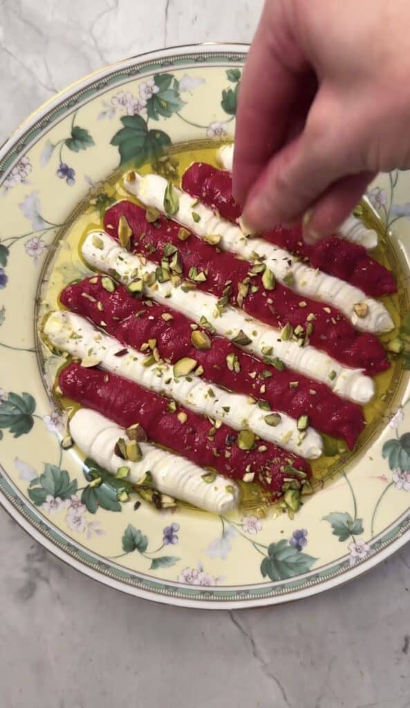 A hand sprinkles chopped pistachios over a plate of Beet & Feta Dip, featuring alternating red and white stripes, drizzled with olive oil on a floral-patterned plate set on a marble surface.