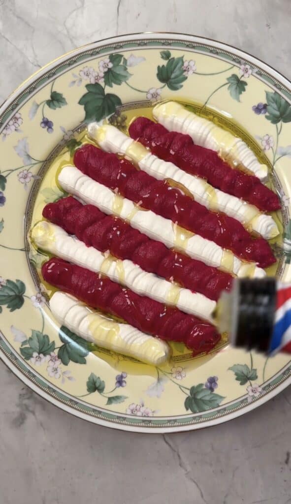 A floral-patterned plate with alternating stripes of Beet & Feta Dip, white cream, and red sauce, drizzled with olive oil, is being topped with balsamic vinegar. The plate sits on a light-colored marble surface.