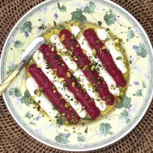 A floral plate on a wicker mat holds rows of creamy white cheese topped with vibrant pink Beet & Feta Dip, chopped pistachios, olive oil, and herbs. A gold-handled knife rests on the plate, with sliced bread visible in the background.