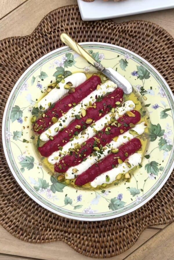 A decorative plate holds a Beet & Feta dip. There are rows of creamy white cheese topped with strips of bright pink beet puree, sprinkled with chopped pistachios and herbs, and drizzled with olive oil. A gold butter knife and slices of bread sit nearby.
