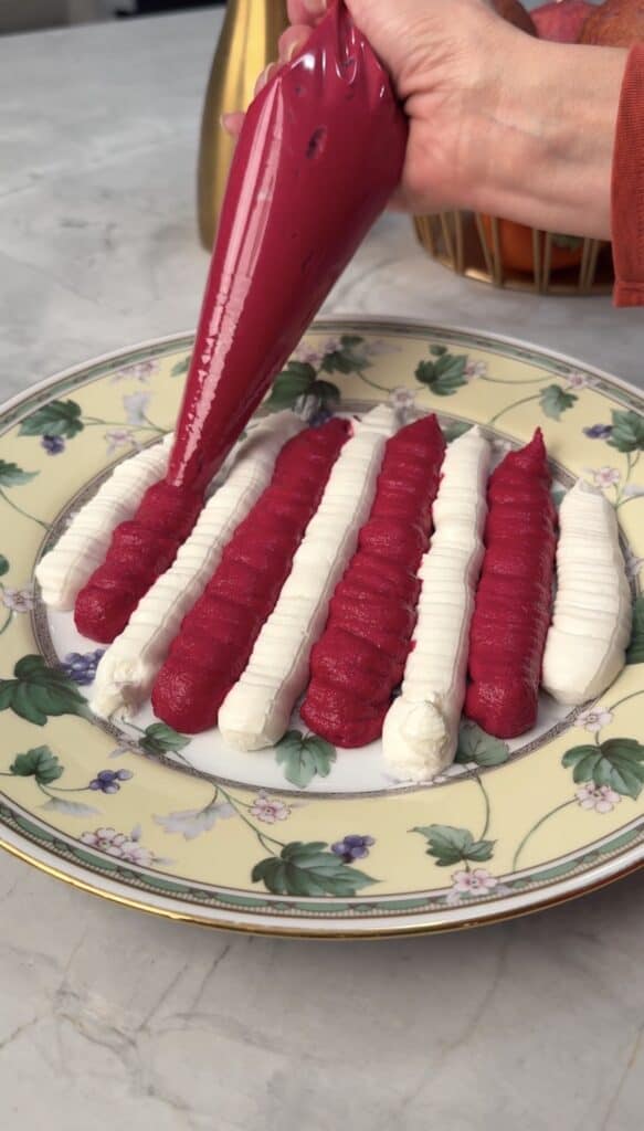 A hand uses a piping bag to add a row of bright pink Beet & Feta Dip next to rows of white cream on a decorative plate with a floral pattern, set on a marble surface.