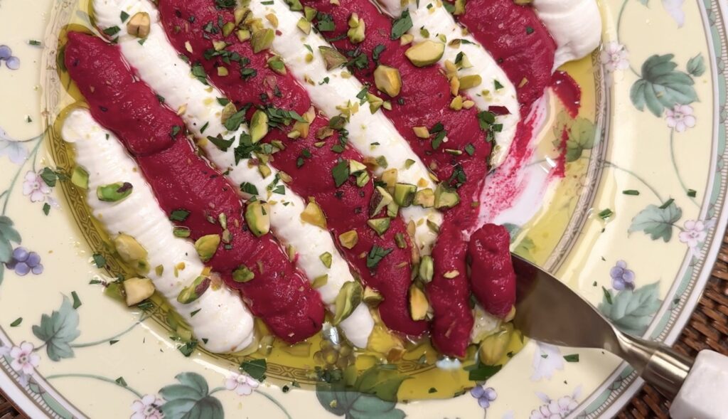 A floral plate with creamy white and bright pink-red Beet & Feta Dip spreads arranged in alternating stripes, topped with chopped pistachios and herbs, and drizzled with olive oil. A spoon rests on the edge of the plate.
