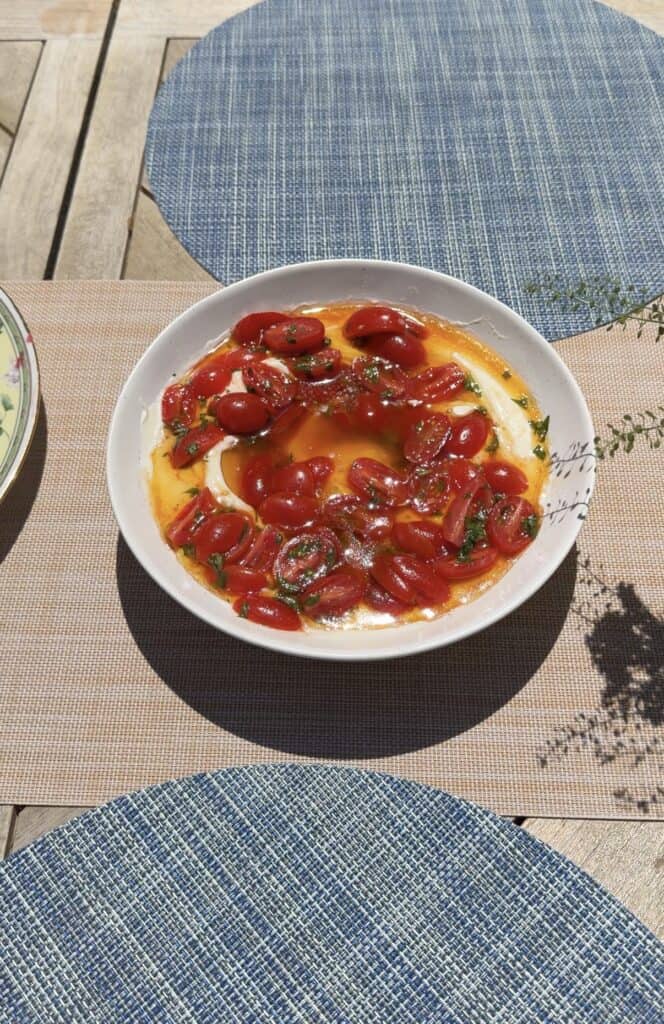 A white bowl filled with bruschetta dip topped with sliced cherry tomatoes, herbs, and oil sits on a woven placemat on a wooden table with blue placemats, bathed in sunlight.