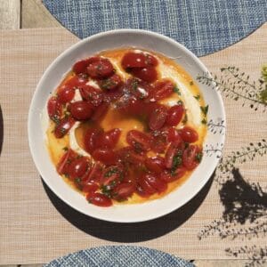 A white plate filled with creamy dip topped with halved cherry tomatoes, fresh herbs, and a drizzle of oil, sitting on a table with blue and beige placemats in sunlight.