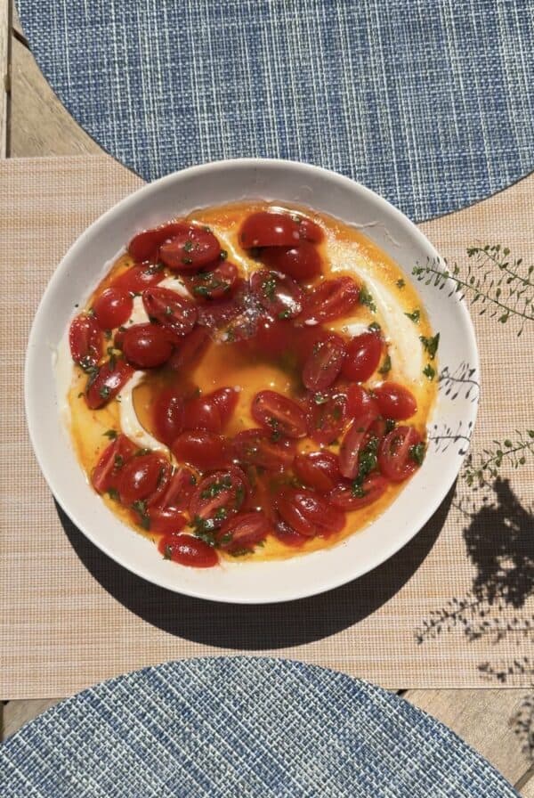 A white plate filled with creamy dip topped with halved cherry tomatoes, fresh herbs, and a drizzle of oil, sitting on a table with blue and beige placemats in sunlight.