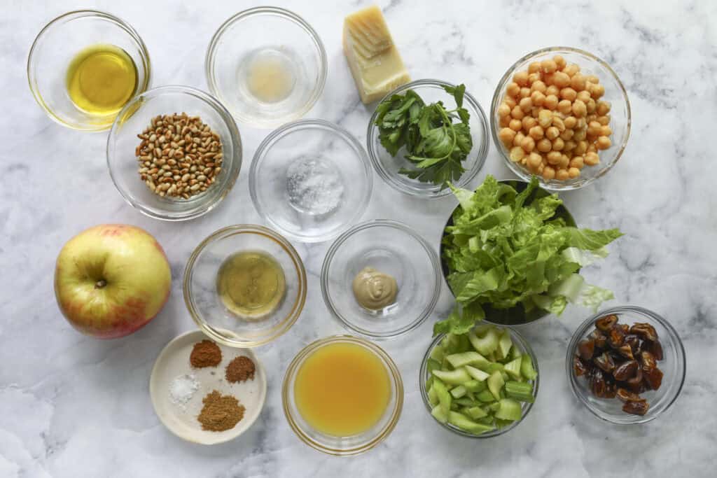 A variety of ingredients for a fresh celery salad are displayed in small glass bowls on a marble surface, including olive oil, chickpeas, lettuce, parsley, pine nuts, apple, dates, grated cheese, spices, vinegar, mustard, and orange juice.