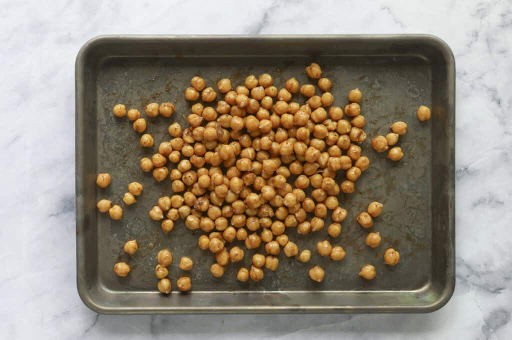 A metal baking tray on a marble surface holds a pile of seasoned chickpeas spaced out and ready for roasting, the perfect crunchy topping for a fresh celery salad.