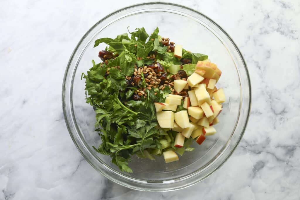 A glass bowl contains chopped parsley, diced apples, sunflower seeds, dates, and crisp celery—all arranged in separate sections on a marble surface before mixing into a vibrant Celery Salad.