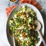 A large oval platter of celery salad with lettuce, apple slices, shaved cheese, crispy chickpeas, and herbs. Wooden salad servers rest in the dish. Nearby are a bowl of chickpeas, a wedge of cheese, and a cheese slicer.