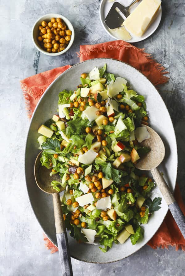 A large oval platter of celery salad with lettuce, apple slices, shaved cheese, crispy chickpeas, and herbs. Wooden salad servers rest in the dish. Nearby are a bowl of chickpeas, a wedge of cheese, and a cheese slicer.