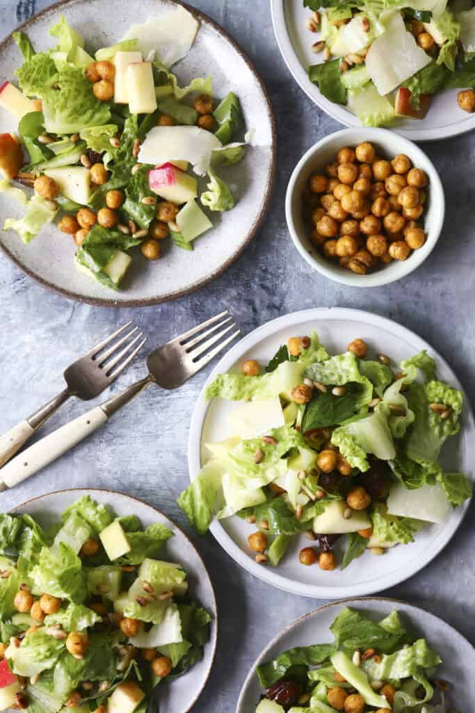 Five plates of green salad with apple slices, parmesan shavings, and crispy roasted chickpeas—this fresh take on celery salad is artfully arranged on a gray surface with a bowl of extra chickpeas and two vintage forks.