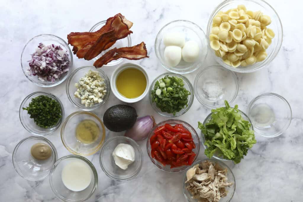 A variety of ingredients for Cobb Pasta Salad in small glass bowls on a marble surface, including cooked pasta, lettuce, grape tomatoes, shredded chicken, avocado, eggs, bacon, chopped herbs, red onion, blue cheese, green onions, and seasonings.