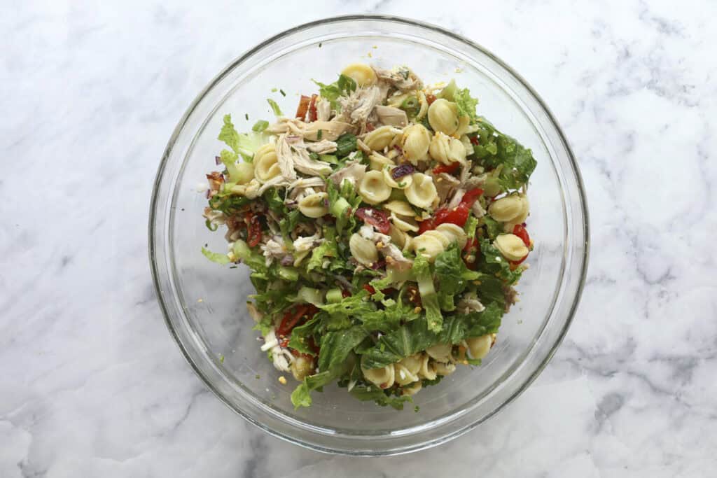 A glass bowl on a marble surface contains a vibrant Cobb Pasta Salad with orecchiette pasta, shredded chicken, lettuce, tomatoes, and fresh herbs.