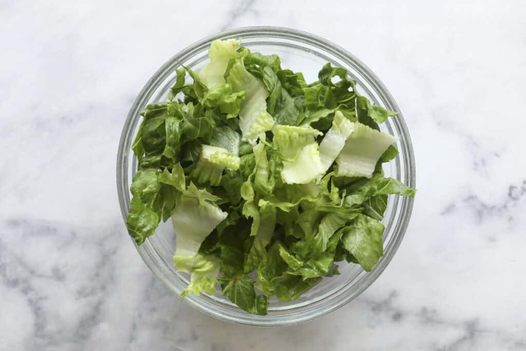 A glass bowl filled with chopped green lettuce sits on a white marble surface, ready to be transformed into a delicious Cobb Pasta Salad.