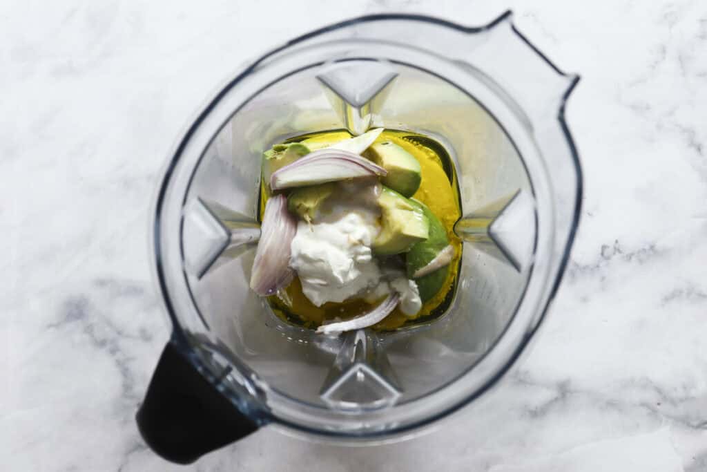 A top-down view of a blender containing ingredients for a fresh Cobb Pasta Salad—avocado, sliced shallot, yogurt, and olive oil—on a white marble surface.