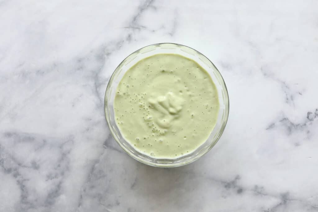 A glass bowl filled with creamy, light green Cobb Pasta Salad dressing sits on a white marble surface.
