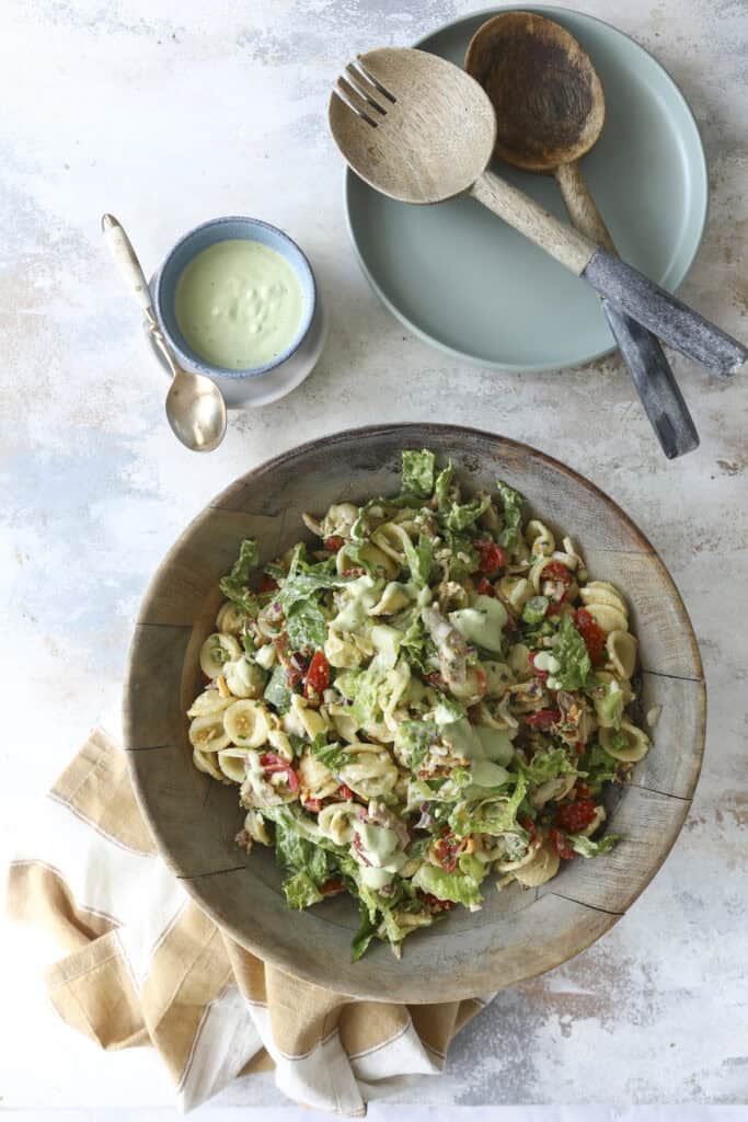 A wooden bowl filled with Cobb Pasta Salad, vegetables, and creamy dressing. Next to it are a plate with wooden serving utensils, a small bowl of green dressing, a spoon, and a folded beige napkin on a light surface.