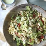 A large wooden bowl filled with Cobb Pasta Salad, featuring lettuce, tomatoes, and a creamy green dressing. A small bowl of extra dressing, a spoon, plates, and wooden salad utensils are nearby on a neutral background.