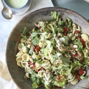 A large wooden bowl filled with Cobb Pasta Salad, featuring lettuce, tomatoes, and a creamy green dressing. A small bowl of extra dressing, a spoon, plates, and wooden salad utensils are nearby on a neutral background.