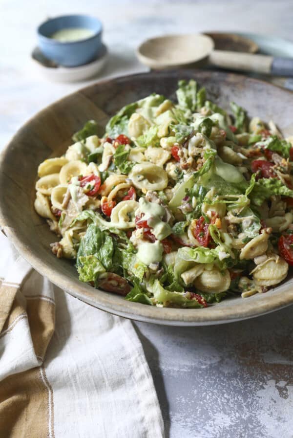 A wooden bowl filled with a fresh Cobb Pasta Salad featuring orecchiette, chopped lettuce, cherry tomatoes, walnuts, and a creamy dressing, with a napkin and small bowl of dressing beside it.