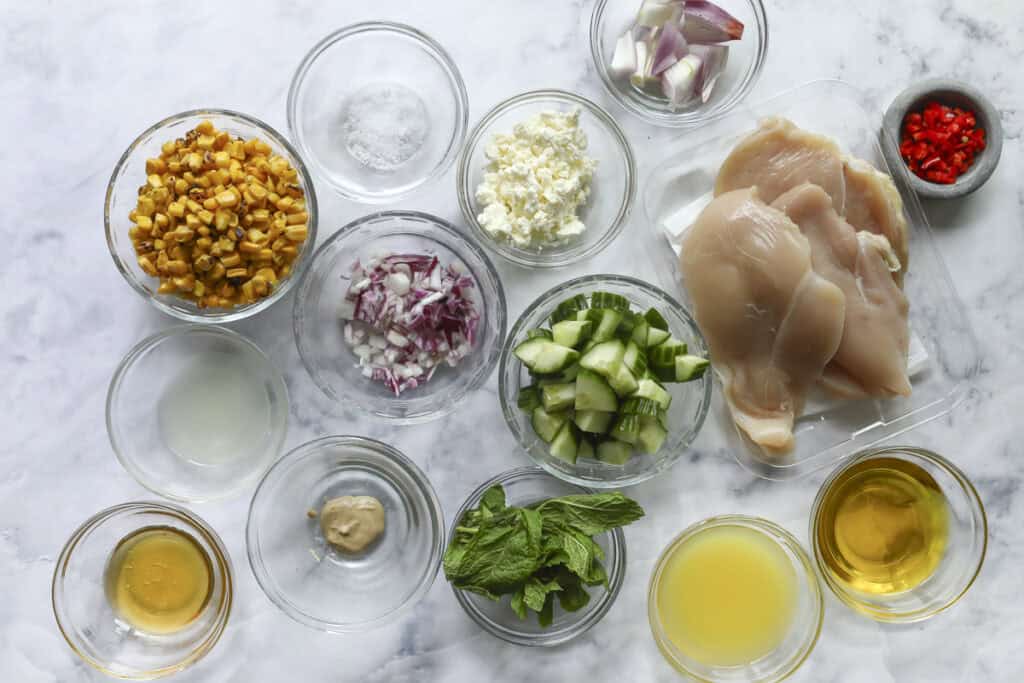 Various ingredients for a Grilled Chicken Summer Salad are arranged in glass bowls on a white surface, including raw chicken breasts, chopped red onion, diced cucumber, roasted corn, mint leaves, feta cheese, lime juice, olive oil, mustard, and red chili.