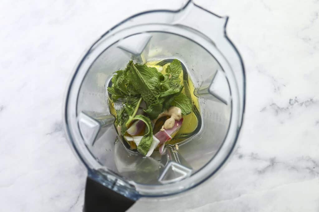 Overhead view of a blender containing fresh mint leaves, red onion pieces, ginger, and a yellow liquid—perfect for dressing a Grilled Chicken Summer Salad—on a white marble surface.
