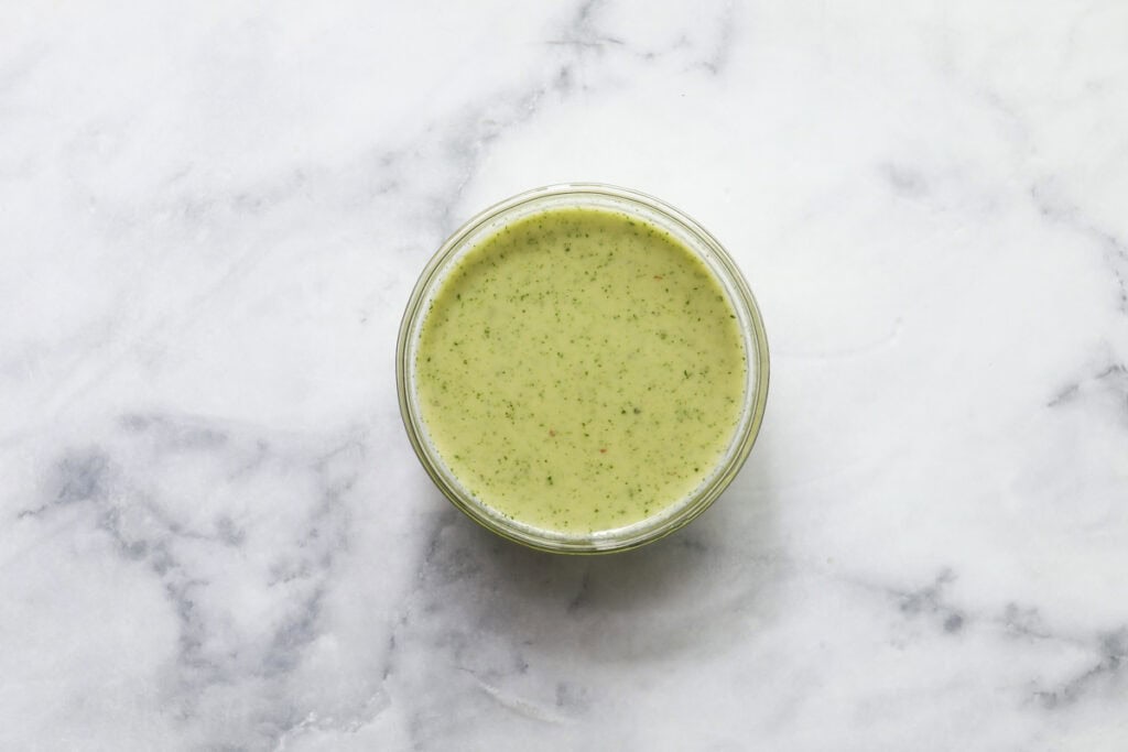 A glass bowl filled with light green sauce, speckled with herbs, sits on a white marble surface—perfect for drizzling over a Grilled Chicken Summer Salad.