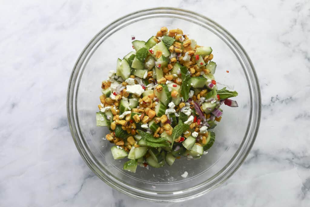 A glass bowl filled with a fresh Grilled Chicken Summer Salad made of chopped cucumber, grilled corn, red onion, feta cheese, mint leaves, and red chili pieces on a white marble surface.