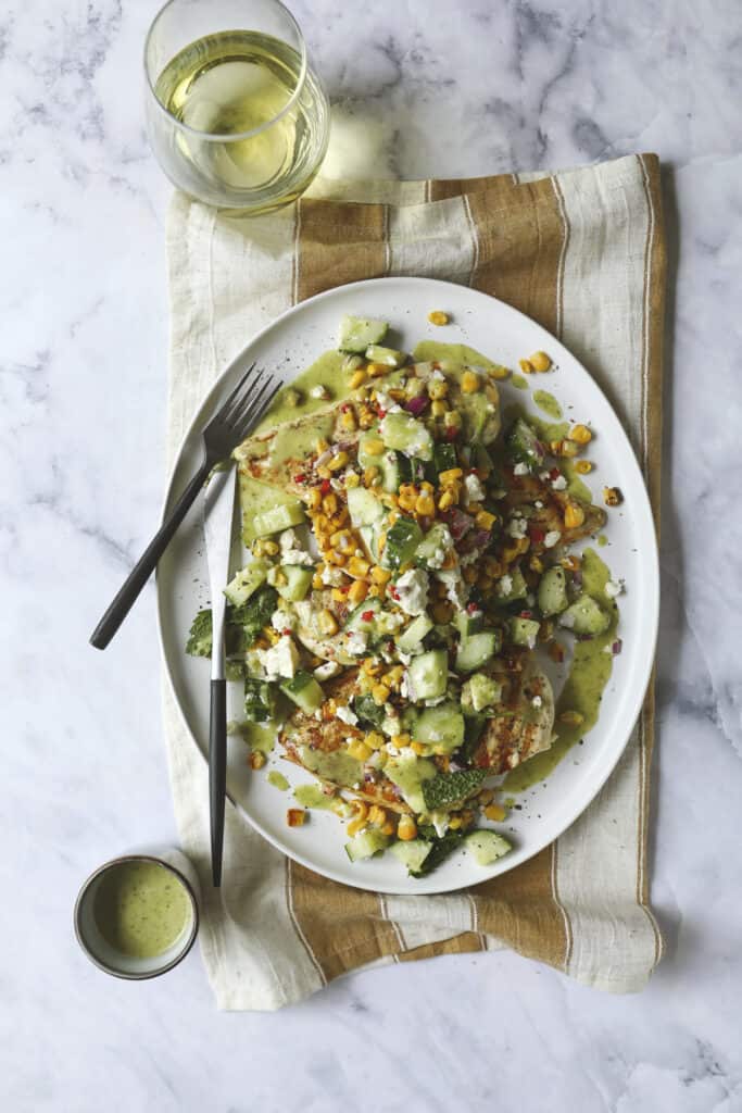 An overhead view of a Grilled Chicken Summer Salad with grilled veggies, chopped cucumber, corn, and creamy dressing on a green sauce, set on a striped napkin. A glass of white wine and a small cup of dressing are beside the plate.
