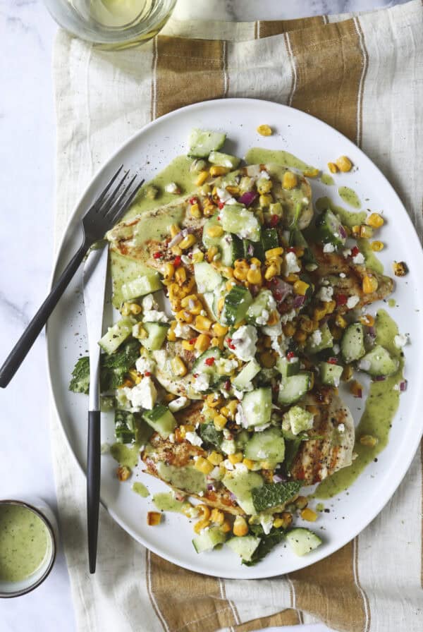 Grilled Chicken Summer Salad features juicy chicken breasts topped with a vibrant mix of corn, cucumber, red onion, herbs, and crumbled cheese, drizzled with green dressing. Served on a white oval plate with cutlery and a glass of white wine nearby.
