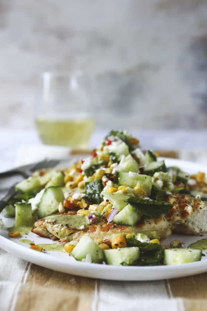 A plate with grilled fish topped with a fresh salad of cucumber, corn, and herbs evokes the vibrant appeal of a Grilled Chicken Summer Salad, set on a striped tablecloth with a glass of white wine in the blurred background.