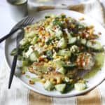 A plate of Grilled Chicken Summer Salad topped with fresh cucumber, corn, herbs, and green dressing sits on a striped cloth napkin with a fork and knife, accompanied by a glass of white wine in the background.