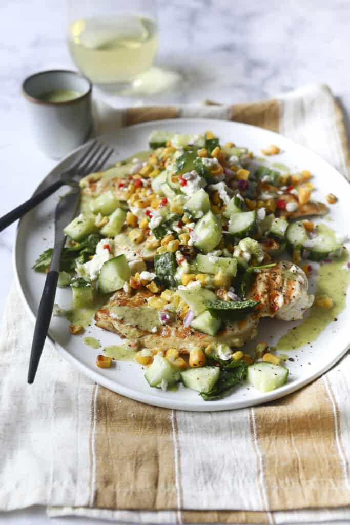 A plate of Grilled Chicken Summer Salad topped with fresh cucumber, corn, herbs, and green dressing sits on a striped cloth napkin with a fork and knife, accompanied by a glass of white wine in the background.