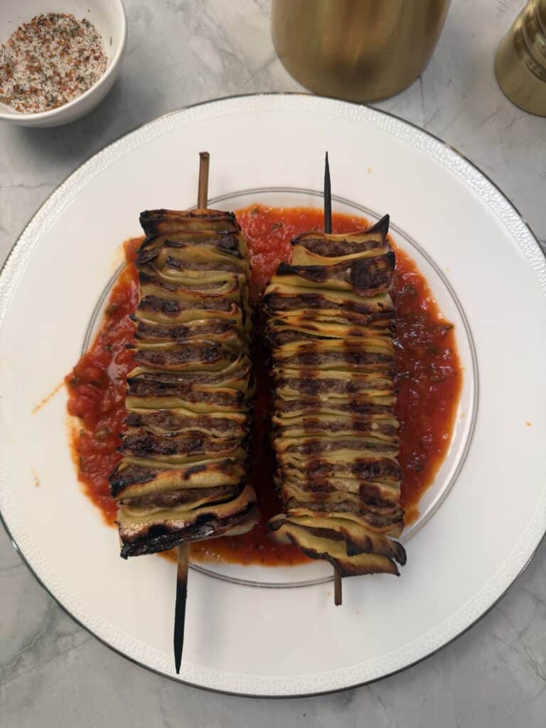 Two grilled lasagna skewers of thinly sliced pasta sheets and meat are stacked on a white plate over red tomato sauce, reminiscent of lasagna on the grill. A bowl of seasoning and a golden container appear in the background.