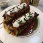 Two grilled lasagna skewers —are topped with tomato sauce, creamy white sauce, and chopped herbs, served on a white plate with a pool of oil and tomato sauce beside gold salt and pepper shakers.