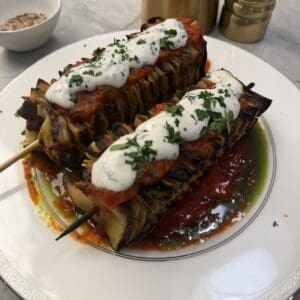 Two grilled lasagna skewers —are topped with tomato sauce, creamy white sauce, and chopped herbs, served on a white plate with a pool of oil and tomato sauce beside gold salt and pepper shakers.