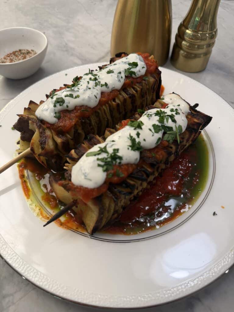 Two grilled lasagna skewers —are topped with tomato sauce, creamy white sauce, and chopped herbs, served on a white plate with a pool of oil and tomato sauce beside gold salt and pepper shakers.