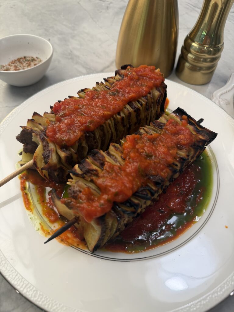 Two skewers of grilled, thinly sliced potatoes are served on a white plate, topped with bright red tomato sauce and green oil—offering flavors reminiscent of lasagna on the grill. Gold salt and pepper shakers and a small bowl of seasoning sit in the background.