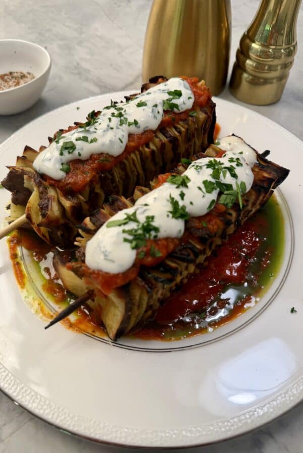 Two grilled lasagna skewers topped with tomato sauce, white creamy sauce, and chopped herbs are served on a white plate with a pool of red and green sauce—bringing the flavors of grilled lasagna to every bite.