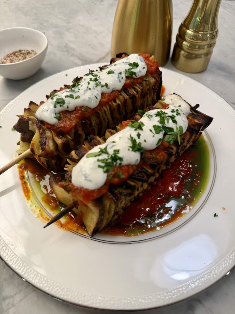 Two grilled vegetable skewers topped with tomato sauce, white creamy sauce, and chopped herbs are served on a white plate with a pool of red and green sauce—bringing the flavors of grilled lasagna to every bite.
