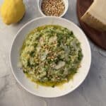 A bowl of vibrant spring risotto garnished with pine nuts, shaved cheese, and herbs sits on a marble surface. Nearby are a lemon, extra pine nuts, and a wedge of cheese on a wooden board.
