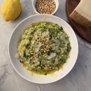 A bowl of vibrant spring risotto garnished with pine nuts, shaved cheese, and herbs sits on a marble surface. Nearby are a lemon, extra pine nuts, and a wedge of cheese on a wooden board.