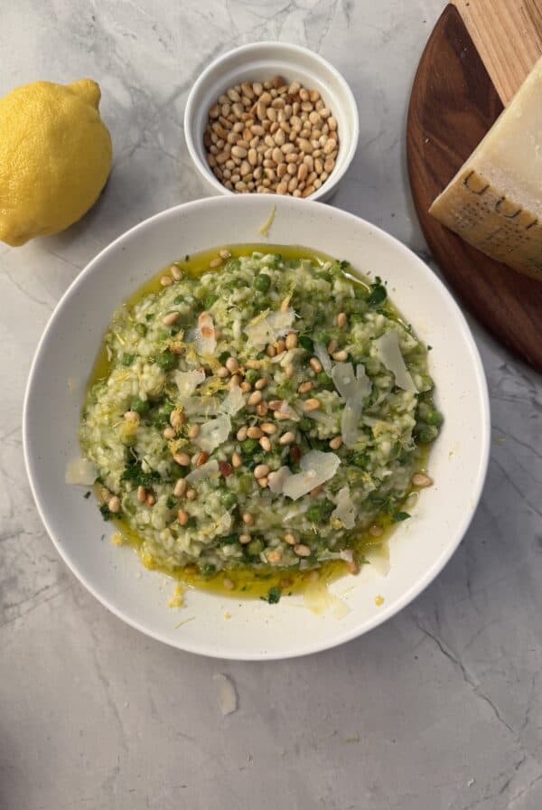 A bowl of vibrant spring risotto garnished with pine nuts, shaved cheese, and herbs sits on a marble surface. Nearby are a lemon, extra pine nuts, and a wedge of cheese on a wooden board.