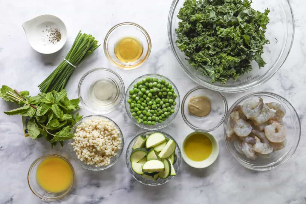 Bowls of fresh ingredients on a marble surface, including kale, peas, zucchini, shrimp, mint, chives, couscous, olive oil, lemon juice, mustard, and seasonings arranged neatly for cooking.