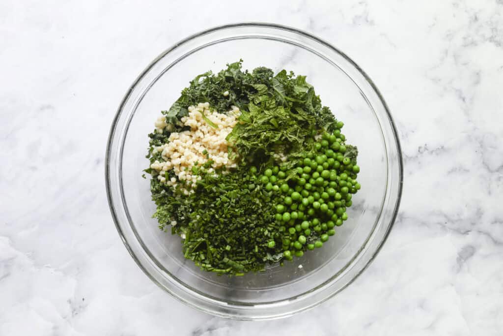 A glass bowl on a marble surface containing separated piles of cooked couscous, chopped greens, fresh mint, parsley, chives, and green peas, ready to be mixed.